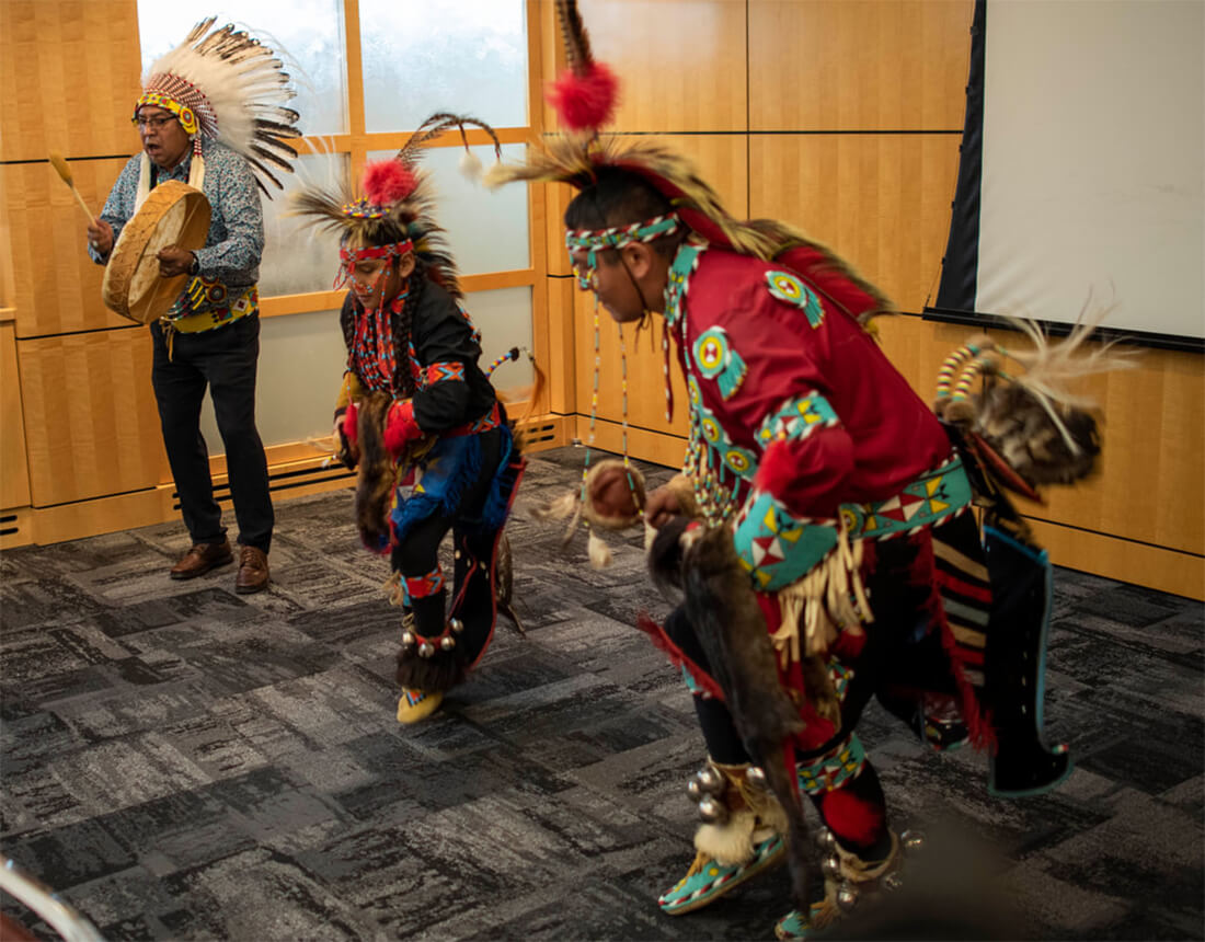 Treffery Deerfoot playing an Indigenous hide drum and 2 traditional indigenous dancers performing in conference room.