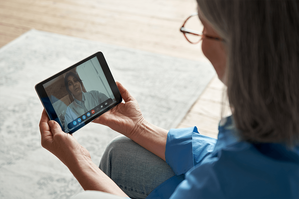 Over the shoulder photo of a seated Woman chatting with health provider on tablet in living room