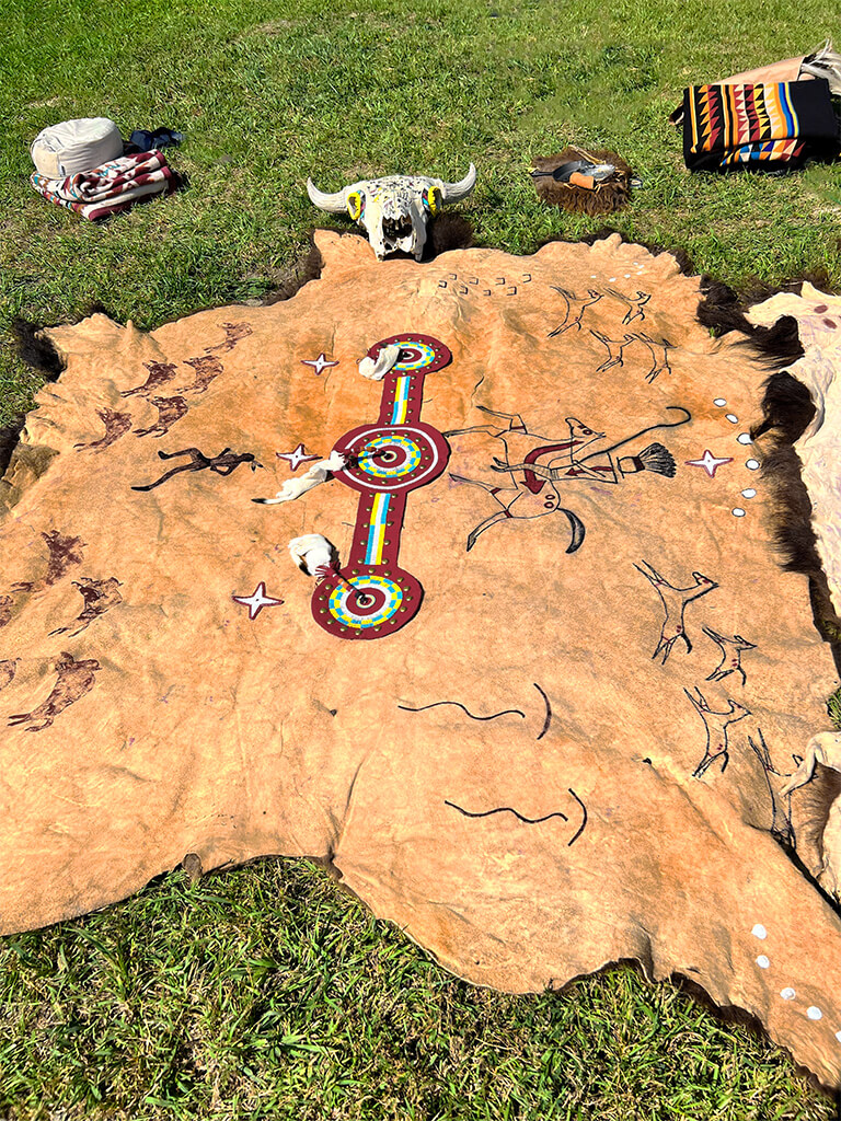 Photo of traditional Deerfoot robe made from animal hide decorated with illustrations of animals and warriors. The robe is laying on grass with animal skull at end of the robe. Other assorted blankets and ceremonial items are near the skull at the far end of the blanket