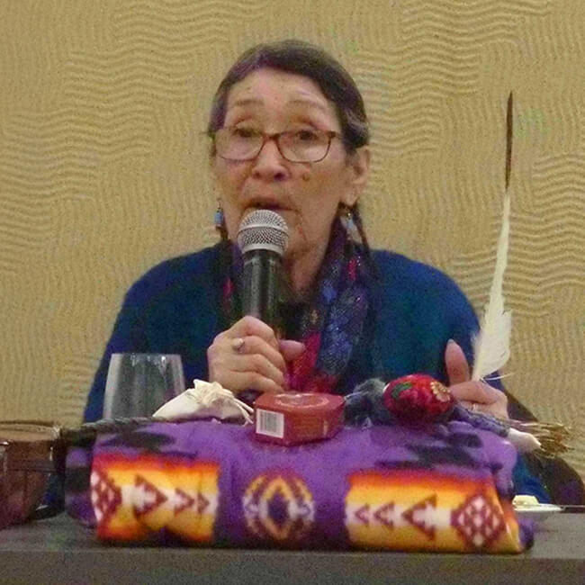 Photo of Elder Doreen Moosepayo sitting at conference table holding an Eagle feather with traditional blanket in front of her while she is speaking into a microphone