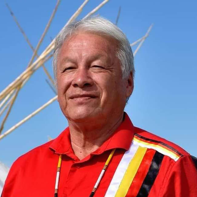 Headshot photo of Elder Gordon Gladue against a clear blue sky. He is wearing a traditional red ribbon shirt with Tipi poles in background.