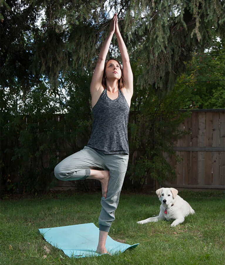 Woman in back yard wearing athletic clothing practicing yoga on a mat with her dog laying on the grass in the background